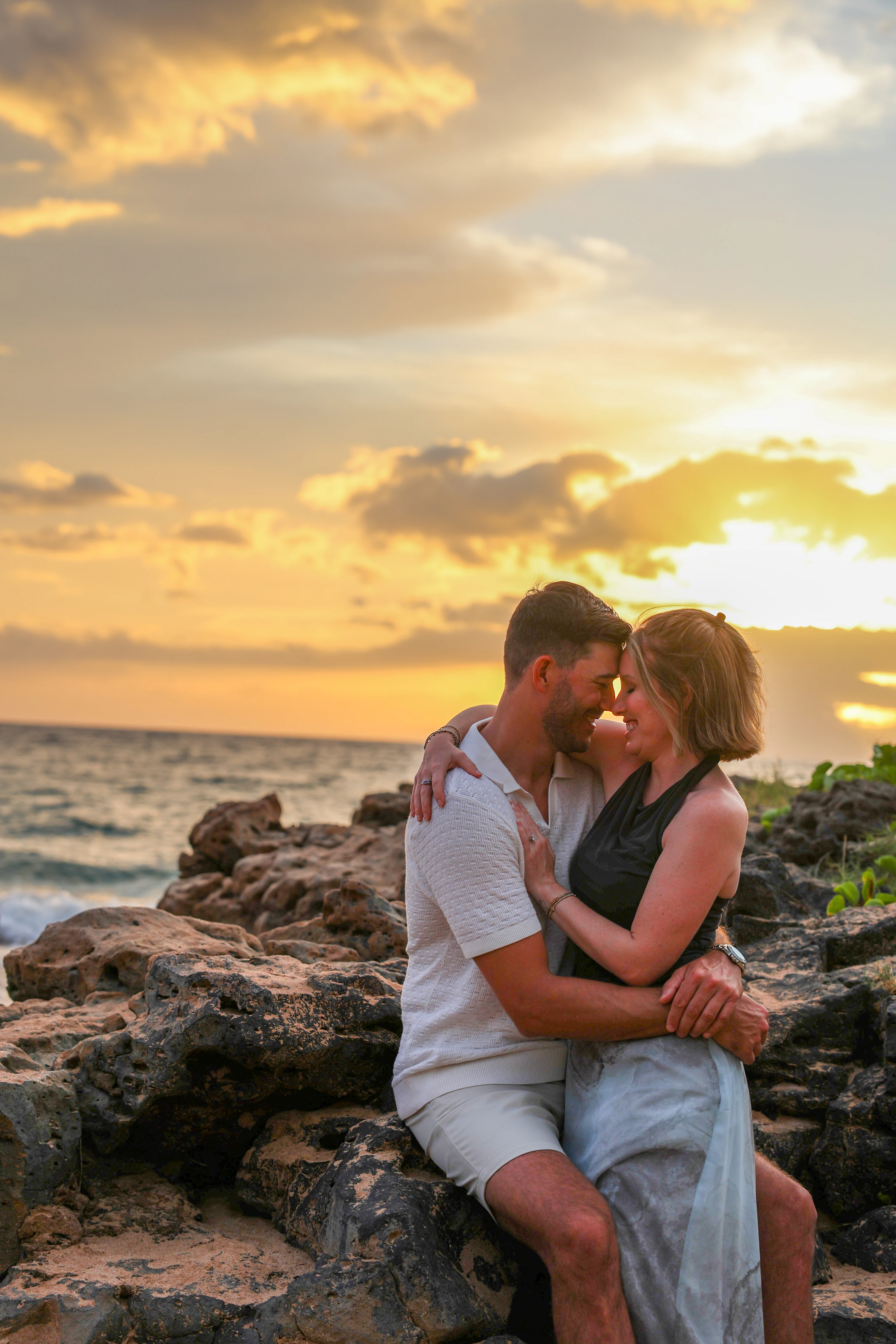 Couple sitting close together on lava rock beneath a golden sky