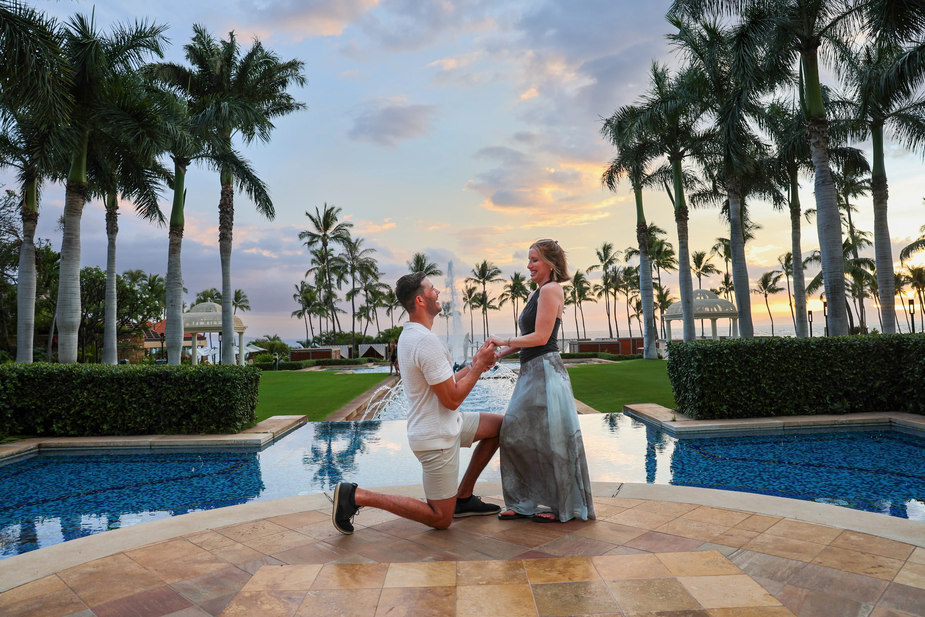 Person proposing beside a resort pool and palms at sunset