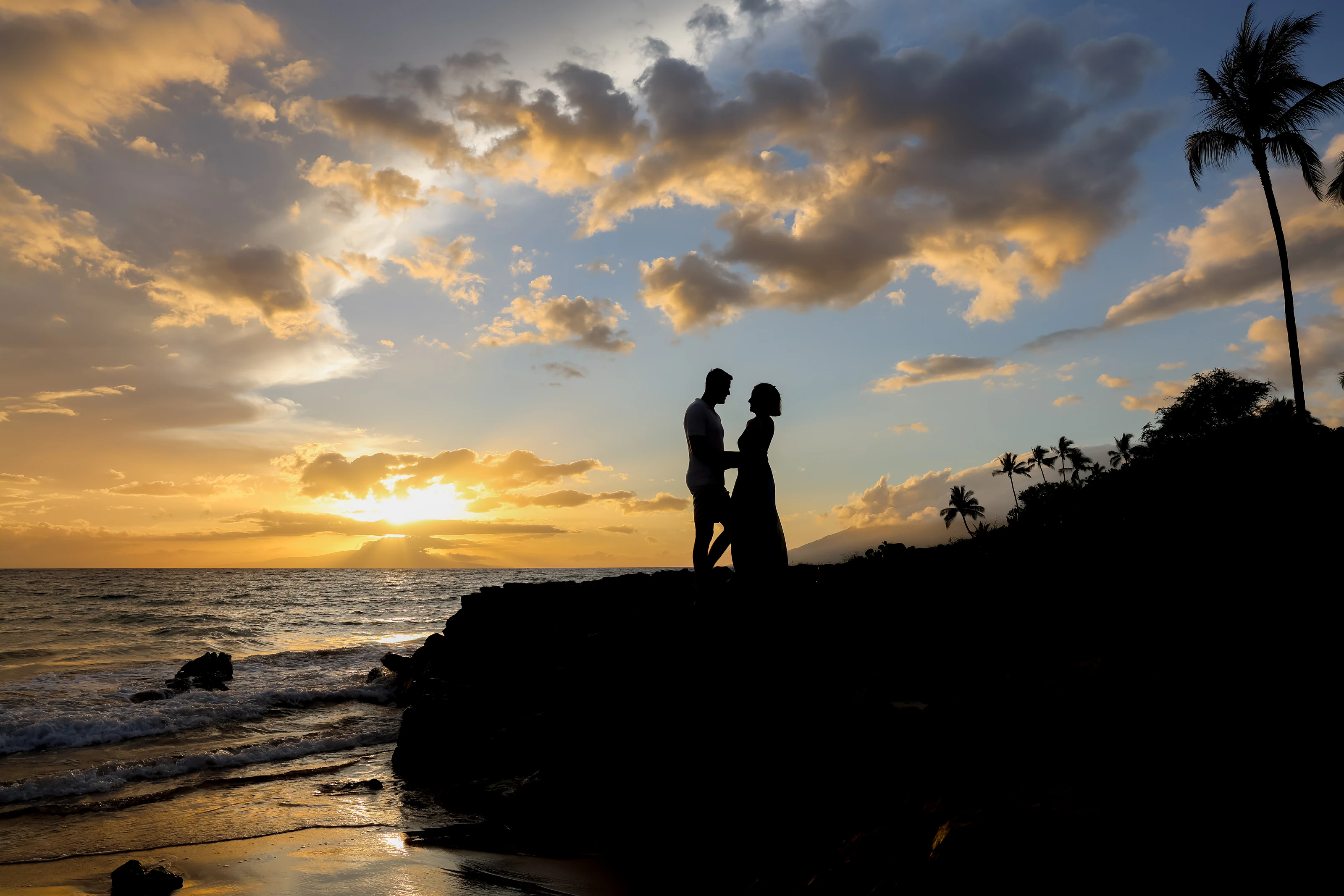 Couple standing in silhouette on lava rock above the ocean beneath golden clouds