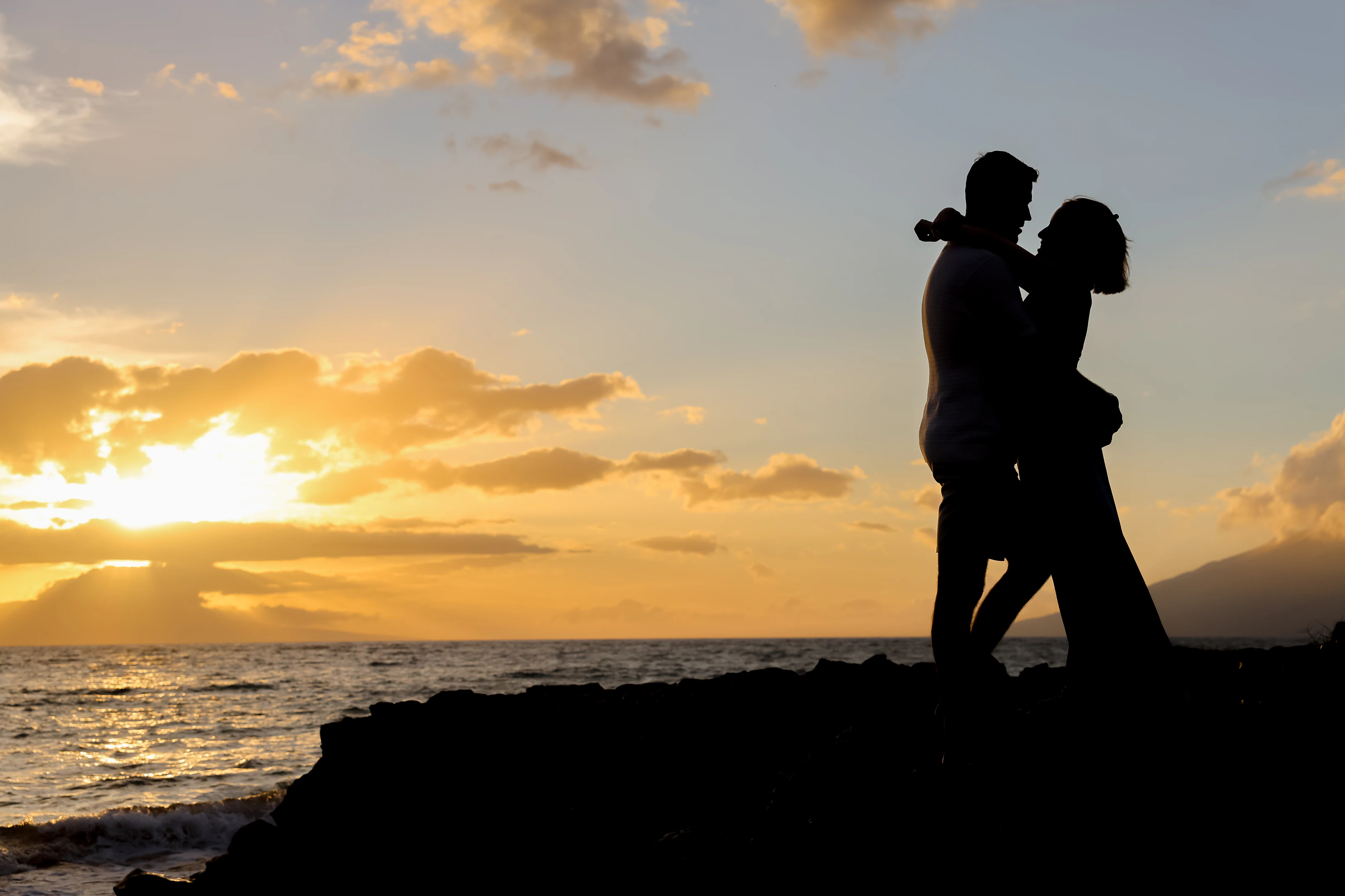 Silhouetted couple embracing on a rocky Maui shoreline at sunset