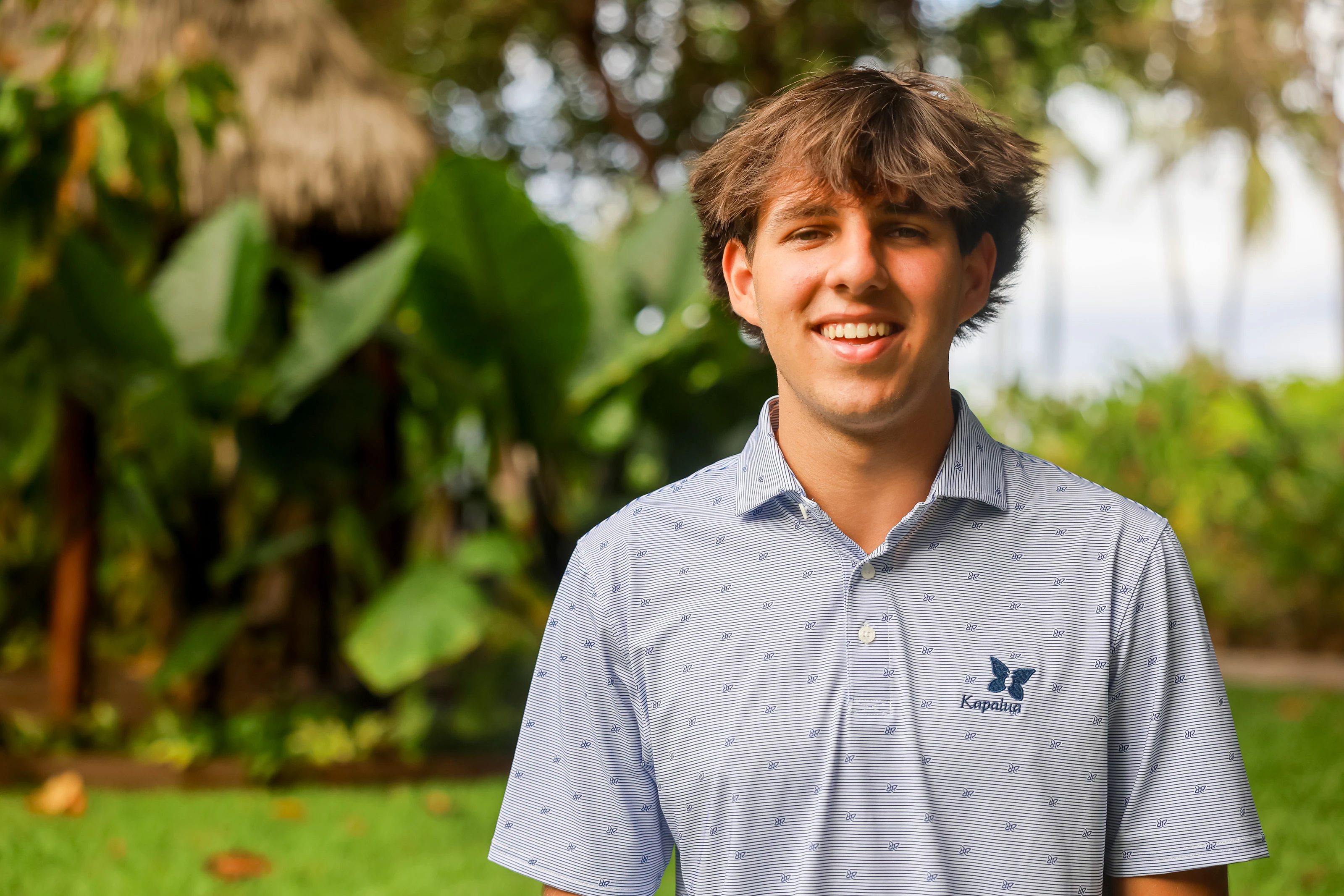 Smiling man in a patterned shirt framed by a sunlit tropical garden