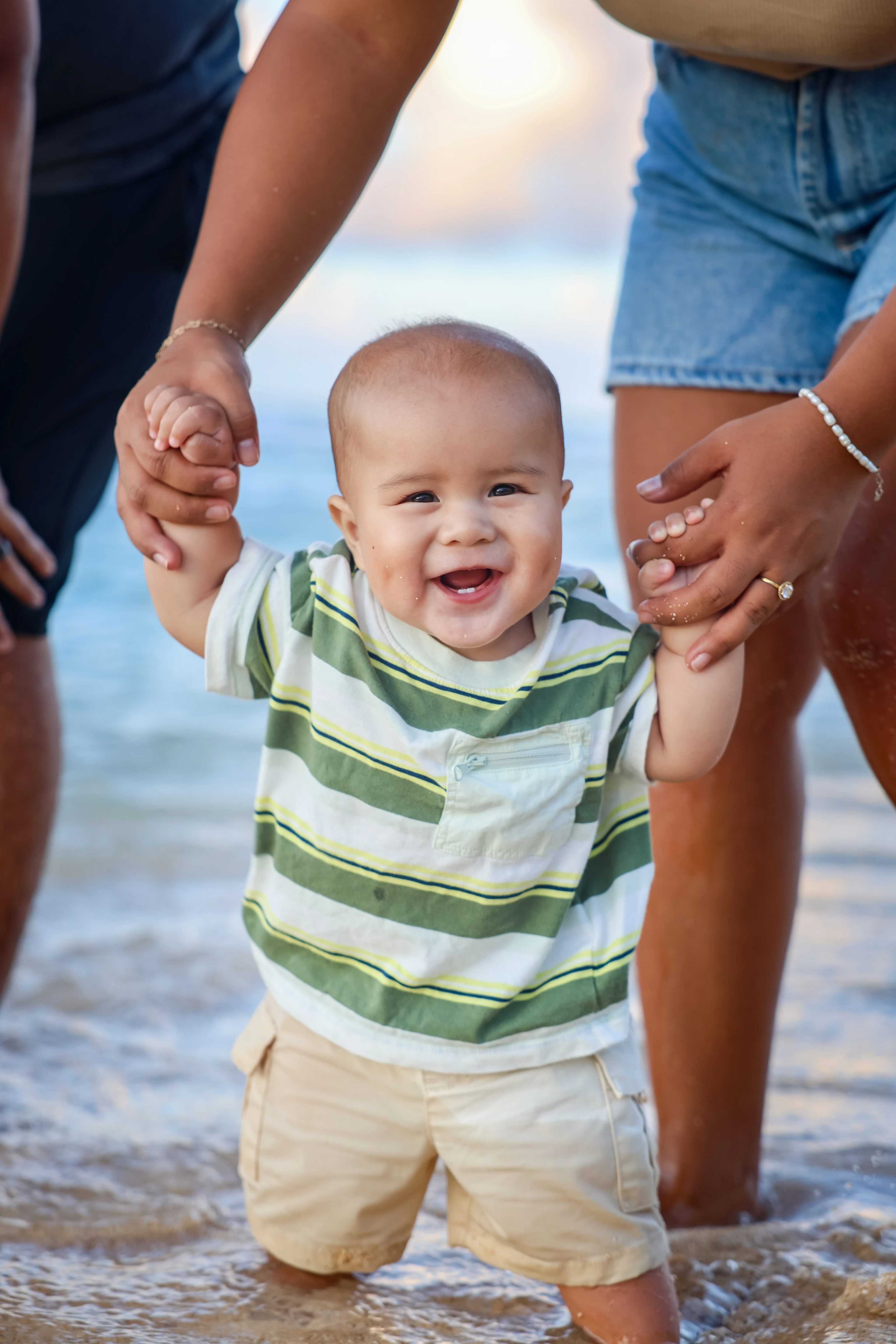 Smiling baby holding both parents’ hands in the shallow ocean water