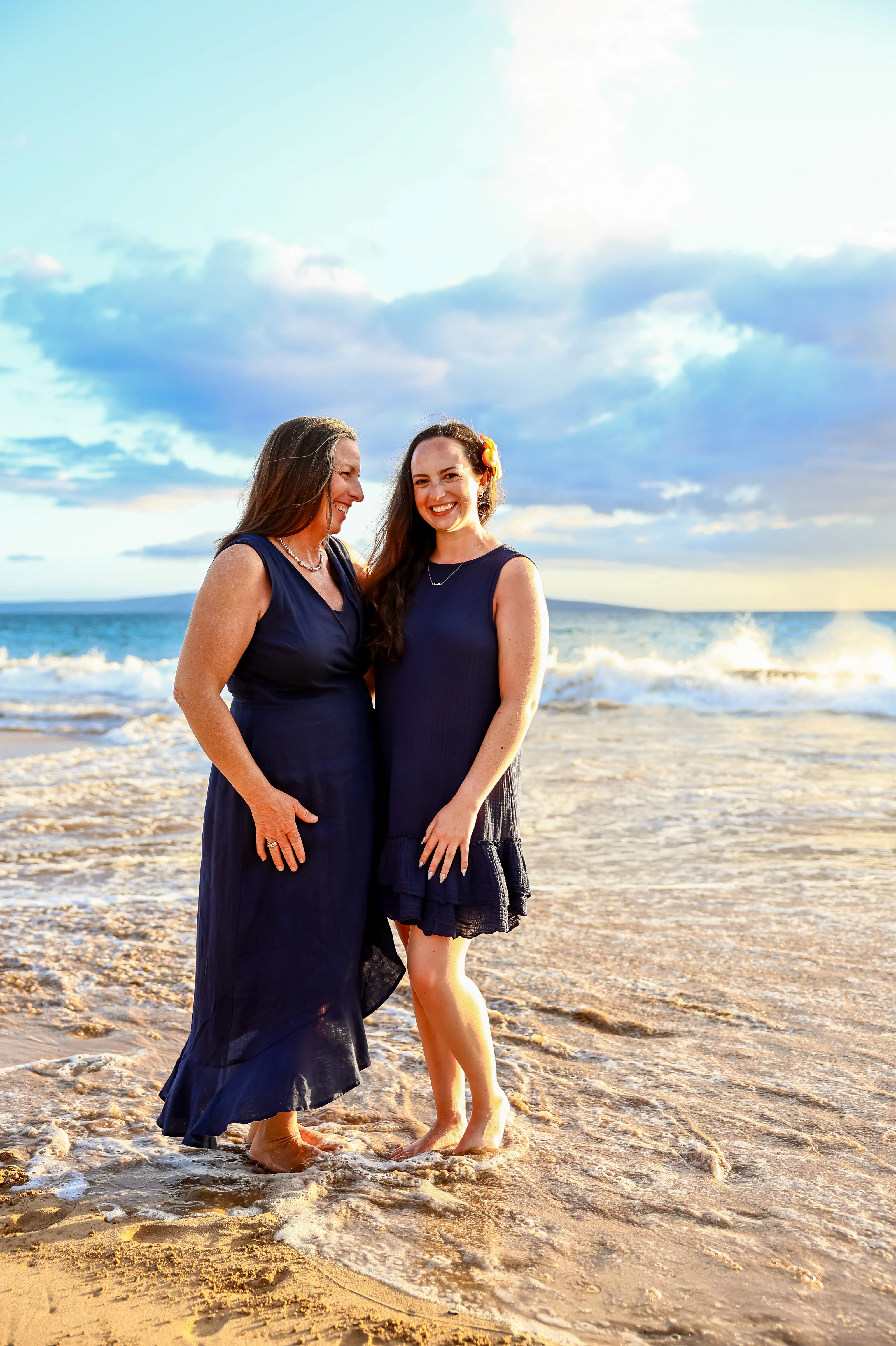 Two women in navy standing together at the waterline during golden hour