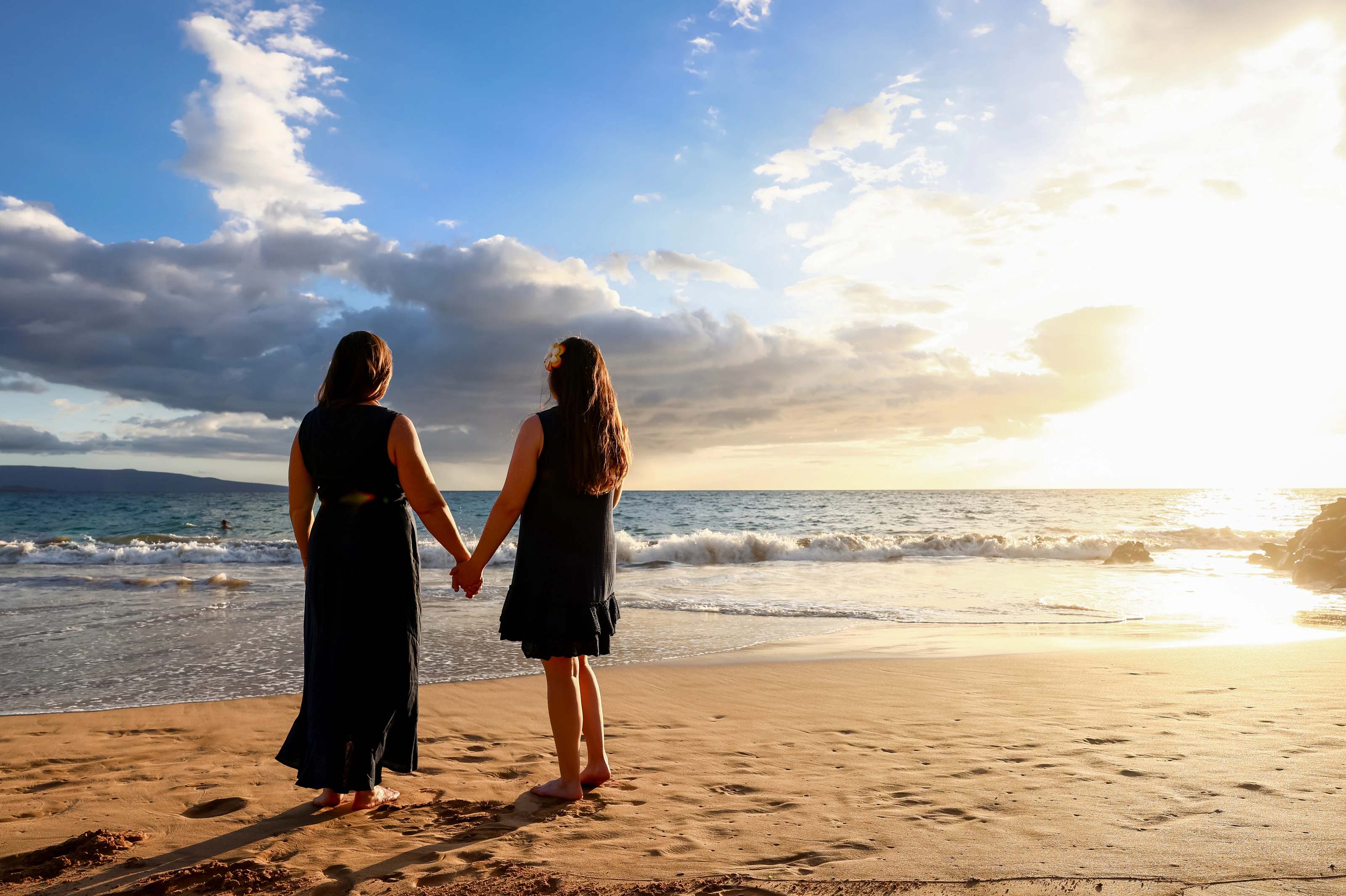 Two women holding hands and looking toward a Maui sunset from the beach