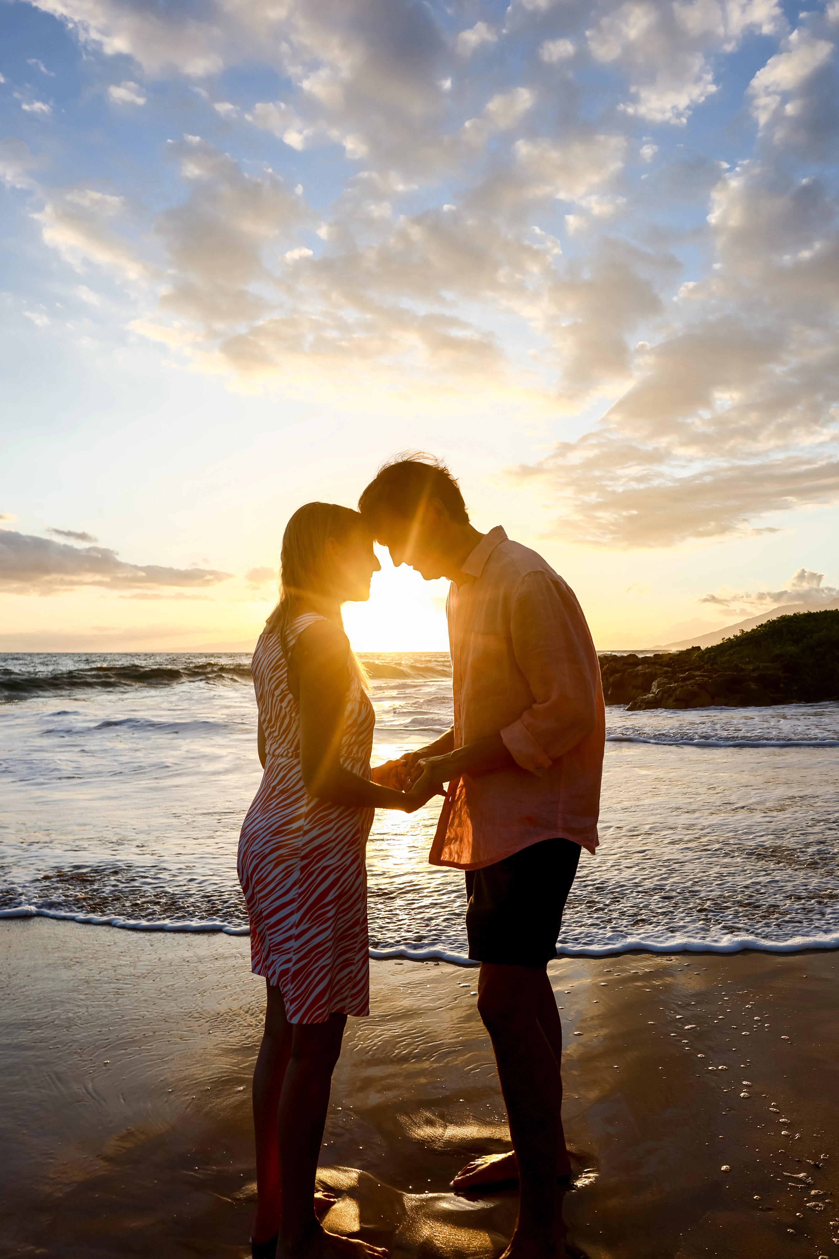 Couple holding hands and touching foreheads at the waterline beneath a wide sunset sky