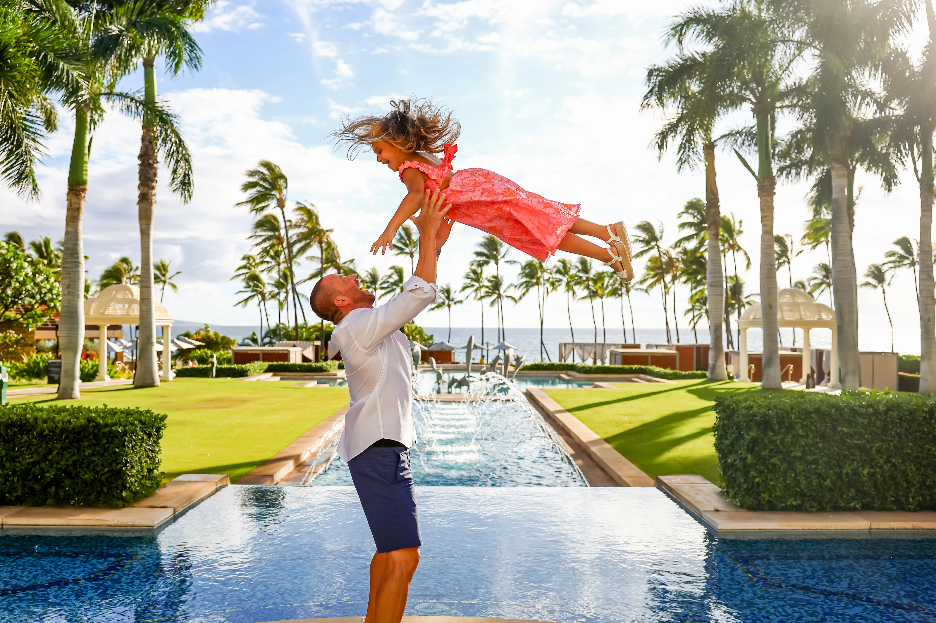 Parent lifting a child high beside a resort pool and palms