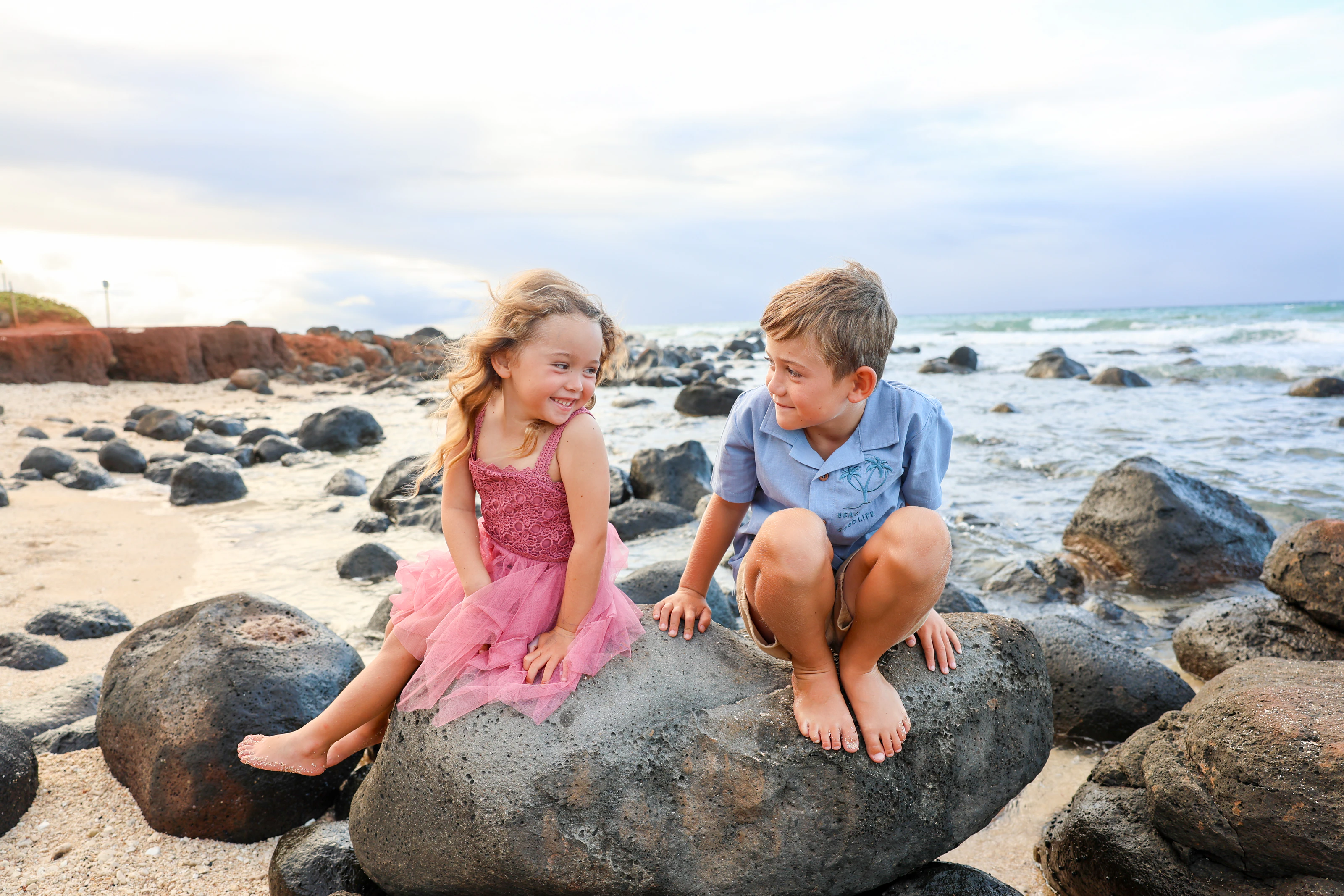 Two children smiling together on lava rocks by the ocean